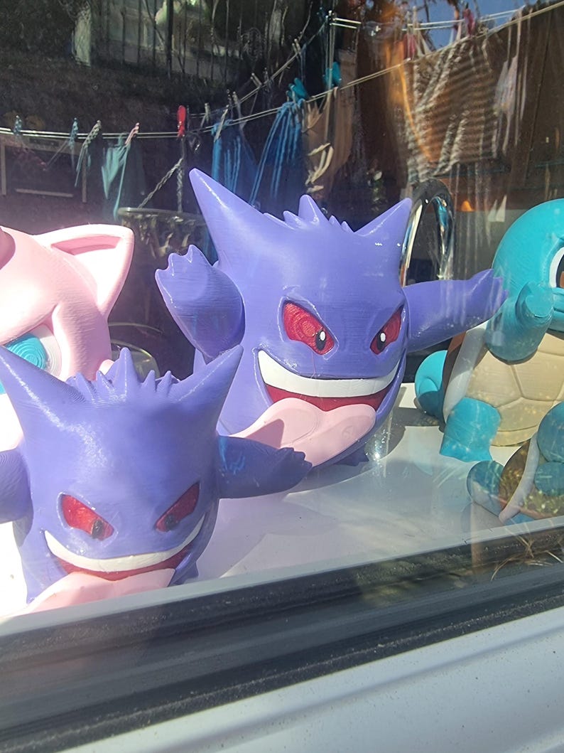 May include: Two purple Gengar figures with pointed ears, red eyes, and wide grins showing white teeth and pink tongues. To the left is a light pink figure with large ears, and to the right is a light blue figure with a tan shell on its back.