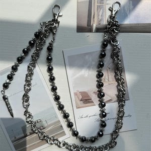 May include: Two silver-toned metal wallet chains with black bead accents and lobster claw clasps. One chain features a spike charm. The chains are displayed on a white surface with printed cards.