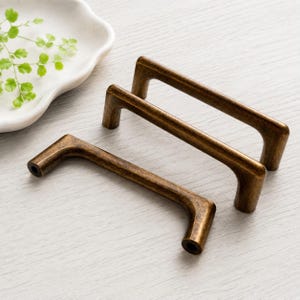 May include: Three bronze-colored cabinet pulls with a rectangular design. The pulls are arranged diagonally, showcasing their curved, angled legs and cylindrical ends. The hardware has a distressed finish, suggesting an antique or vintage aesthetic.