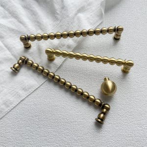 Op de afbeelding: Drie antieke messing kastgrepen en een knop. De grepen hebben een reeks kleine, ronde kralen. De hardware heeft een warme, gouden tint en is gerangschikt op een wit oppervlak.