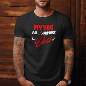 Op de afbeelding: Zwart T-shirt met de tekst "MY EGO WILL SURPRISE YOU" in rode en witte letters. Het woord "You" is in een cursief rood lettertype geschreven. Het shirt heeft een ronde hals.