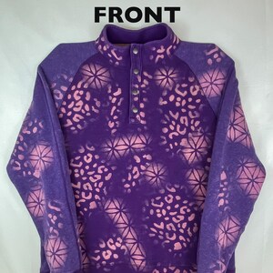 May include: A purple pullover sweatshirt with a high collar and three buttons. The sweatshirt features a pattern of pink floral and leopard print designs. The word "FRONT" is printed at the top of the image.