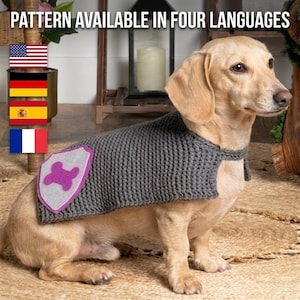May include: A tan dachshund wearing a gray knitted cape with a felt shield featuring a purple bone design. The text "PATTERN AVAILABLE IN FOUR LANGUAGES" is at the top, with flags of the USA, Germany, Spain, and France.