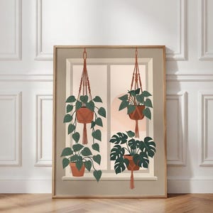 May include: A framed art print featuring a window scene with hanging plants. The illustration shows three potted plants suspended by macrame hangers. The plants have green leaves and are set against a neutral background with a warm, peachy glow.