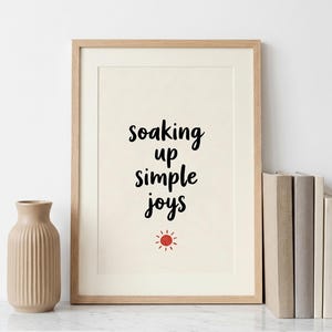May include: A framed print with the words "soaking up simple joys" in black script, with a small red sun graphic. A beige vase and a stack of books are on the left and right.