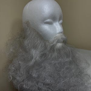 May include: A white foam mannequin head with a long, gray, curly beard and mustache attached. The beard is full and covers the neck and chest area. The mannequin is set against a neutral background.
