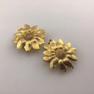 May include: Two gold-colored flower-shaped clip-on earrings. Each earring features detailed petals and a textured center, resembling a sunflower. The earrings are set against a white background.