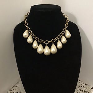 May include: A gold-toned chain necklace with large, pear-shaped, off-white faux pearls. The necklace is displayed on a black mannequin, showcasing its design and elegance. The pearls hang from the chain, creating a cascading effect.