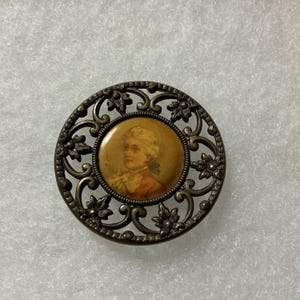 May include: A round brooch featuring an antique-style portrait of a person with light hair in a tan and brown color palette. The central image is encased in a dark metallic frame with an intricate openwork design of swirling scrolls and leaves.