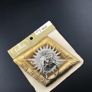 May include: A gold-toned diamond-shaped button with a textured surface and a central silver-toned sunburst emblem featuring a lion head holding a ring. The button is attached to a beige card labeled La Mode Buttons with handwritten text 8.00 $6.00.