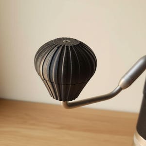 Könnte beinhalten: Ein schwarzer, gerippter, kugelförmiger Griffknopf, der an einem metallischen, abgewinkelten Kurbelarm befestigt ist. Der Knopf hat eine texturierte, vertikal geriffelte Oberfläche mit einer kleinen, mittigen Sechskantschraube oben.