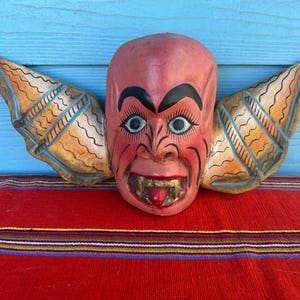 Op de afbeelding: Een handgesneden houten masker dat is geschilderd in rood, oranje en blauw, en een demon met vleugels voorstelt. Het masker heeft een woeste uitdrukking met scherpe tanden en een uitstekende tong.