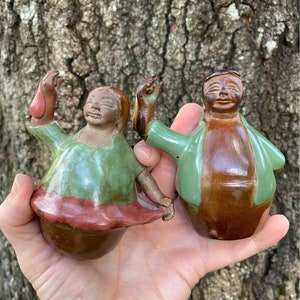 Puede incluir: Dos figuritas de cerámica, una con un esmalte verde y marrón y la otra con un esmalte verde, marrón y rojo. Las figuritas son estilizadas y tienen rasgos simples.