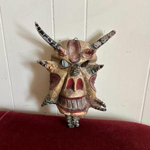 May include: A carved wooden mask featuring a tan base with red and black painted accents. The mask has two large upward-curving horns, several smaller protruding spikes, hollow eye sockets, a prominent nose, and a wide, toothy grin.