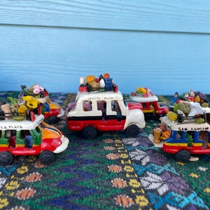 May include: Five colourful ceramic toy buses with various destinations written on the sides, including "La Plata", "San Agustin", and "Pamplitas, Huila". The buses are decorated with flowers, fruit, and other details, and are arranged on a patterned rug.