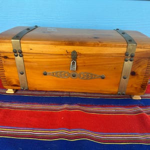 May include: A wooden chest with brass accents, including corner brackets and decorative embellishments. The chest is secured with a padlock and sits on small, light-colored feet. The chest is on a striped red, blue, and yellow textile.
