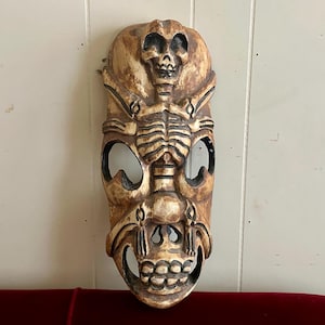 May include: A wooden mask with a carved skull and skeleton design. The mask is painted in shades of brown and white.