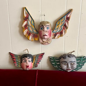 Op de afbeelding: Drie handgeschilderde houten engelenmaskers met vleugels. Het bovenste masker heeft een blond hoofd en kleurrijke vleugels. De andere twee maskers hebben donker haar en rode, groene en witte vleugels. De maskers hangen aan een witte muur.