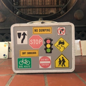 Puede incluir: Una fiambrera de metal blanca con una superficie gris cubierta de varias se&ntilde;ales de tr&aacute;fico. Las se&ntilde;ales incluyen "No Dumping", "Stop", "Soft Shoulders", "Bike Route", "Do Not Enter", y una se&ntilde;al con un coche y un camino sinuoso.