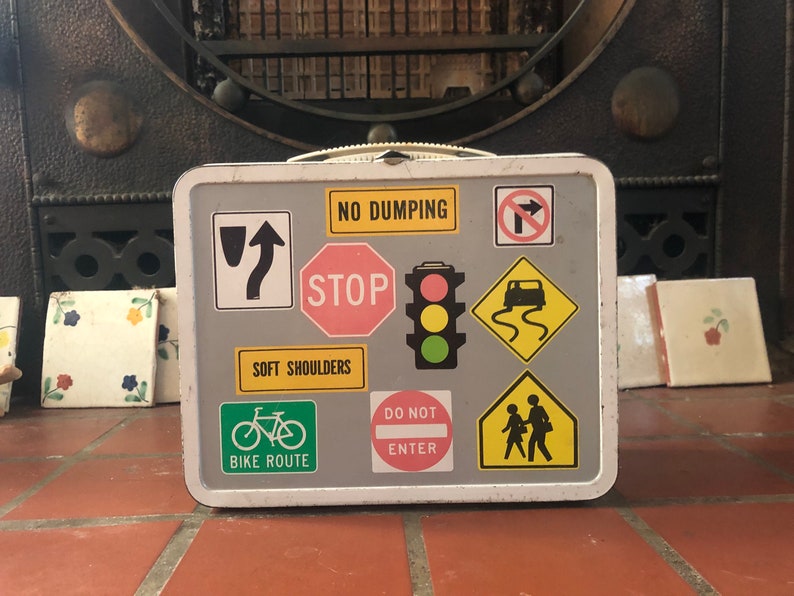 Puede incluir: Una fiambrera de metal blanca con una superficie gris cubierta de varias se&ntilde;ales de tr&aacute;fico. Las se&ntilde;ales incluyen "No Dumping", "Stop", "Soft Shoulders", "Bike Route", "Do Not Enter", y una se&ntilde;al con un coche y un camino sinuoso.