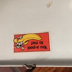 Puede incluir: Una fiambrera de metal blanca con una pegatina de pl&aacute;tano amarilla que dice "You a-peel to me!"