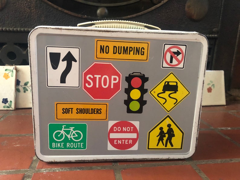 Puede incluir: Una fiambrera de metal con fondo gris y varias se&ntilde;ales de tr&aacute;fico y s&iacute;mbolos. Las se&ntilde;ales incluyen "No Dumping", "Stop", "Soft Shoulders", "Bike Route", "Do Not Enter", y "School Crossing".