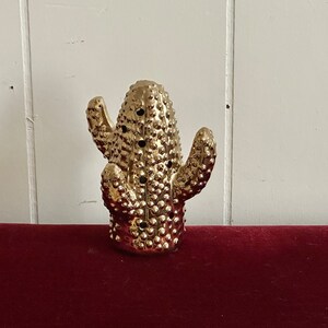 May include: A gold-colored ceramic cactus sculpture with a textured surface. The cactus has multiple arms and is standing on a red surface.