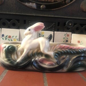 May include: A white ceramic planter shaped like a rabbit with pink ears, standing on a bed of green and black leaves. The planter has a pink and green floral design.