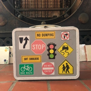Puede incluir: Una fiambrera vintage de metal con fondo gris y varias se&ntilde;ales de tr&aacute;fico y s&iacute;mbolos. Las se&ntilde;ales incluyen "No Dumping", "Stop", "Soft Shoulders", "Bike Route", "Do Not Enter" y una se&ntilde;al de paso de peatones.