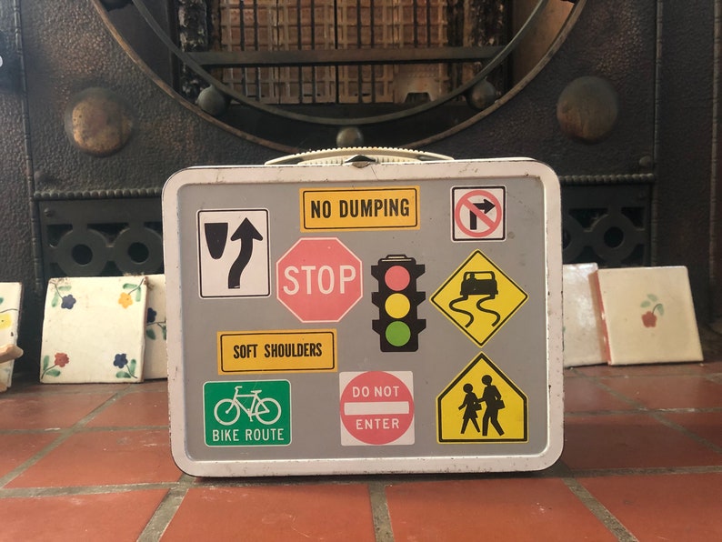 Puede incluir: Una fiambrera vintage de metal con fondo gris y varias se&ntilde;ales de tr&aacute;fico y s&iacute;mbolos. Las se&ntilde;ales incluyen "No Dumping", "Stop", "Soft Shoulders", "Bike Route", "Do Not Enter" y una se&ntilde;al de paso de peatones.