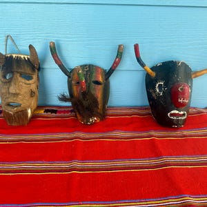 Op de afbeelding: Drie handgemaakte houten maskers met hoorns, tentoongesteld op een rood en gestreept textiel tegen een blauwe achtergrond. De maskers hebben verschillende kleuren en ontwerpen, waaronder een gebeeldhouwd gezicht, geschilderde details en een stierachtig uiterlijk.