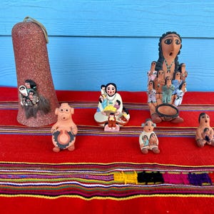 May include: A selection of handmade ceramic figurines, including a tall, textured, reddish-brown bell-shaped piece and several small figures depicting people. The figurines are displayed on a red textile with colourful stripes.