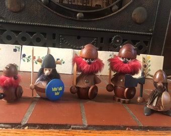 Puede incluir: Cinco figuritas vikingas de madera pintadas en rojo y marrón. Las figuritas llevan cascos y tienen barbas de piel roja. Una figurita sostiene una espada, otra un hacha y otra un escudo con el escudo de armas sueco.