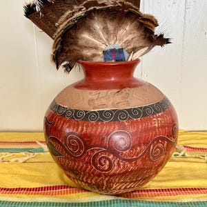 May include: A red clay pot with black and white painted designs. The pot is decorated with swirling patterns and a stylized bird. A fan of brown feathers sits on top of the pot.
