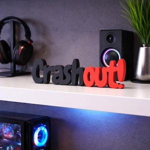Könnte beinhalten: Ein schwarzes und rotes Holzschild mit der Aufschrift "Crashout!". Das Wort "Crash" ist schwarz und "out!" ist rot. Das Schild steht auf einem weißen Regal, im Hintergrund sind ein Computer, ein Lautsprecher und Kopfhörer zu sehen.