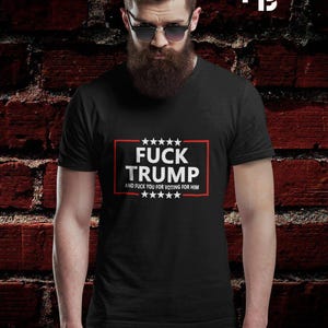 May include: Black t-shirt with the text "FUCK TRUMP" in a white font inside a red rectangle. Below the text is the phrase "AND FUCK YOU FOR VOTING FOR HIM" in white. The background is a red brick wall.