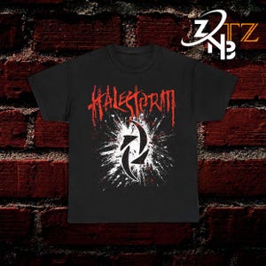 May include: A black short-sleeve t-shirt featuring the red text Halestorm in a jagged font above a white circular graphic with a black abstract symbol in the center, surrounded by white paint splatter effects on the front of the garment.