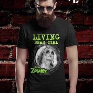 Puede incluir: Camiseta negra con un diseño de una mujer con pelo largo blanco dentro de un círculo oscuro. El texto "LIVING DEAD GIRL" y "ROB ZOMBIE" están impresos en verde.