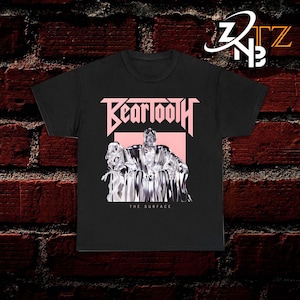 May include: A black short-sleeve t-shirt featuring a graphic print with the text "BEARTOOTH" in light pink above a metallic, chrome-like figure seated on a throne, set against a light pink rectangular background with the text "THE SURFACE" below.