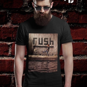 May include: Black t-shirt featuring the band name "RUSH" in a bold font. The graphic shows a person kicking a ball near water. The background is a brick wall. The person in the image is wearing sunglasses.