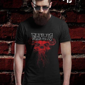 May include: Black t-shirt featuring the band name "DANZIG" in white, above a red skull graphic with horns. The image is set against a red brick wall background.