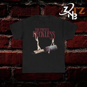 May include: A black t-shirt featuring the text THE PRETTY RECKLESS in dark red lettering above a graphic of a round hairbrush wrapped in white fabric strips. The brush has a blue and red striped barrel and is positioned horizontally in the center.