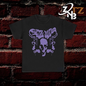 May include: A black short-sleeved t-shirt featuring a purple graphic print on the chest. The design displays a central human skull flanked by two figures, with the text "ELECTRIC WIZARD" arched above in a stylised, flowing font. The shirt has a crew neck.