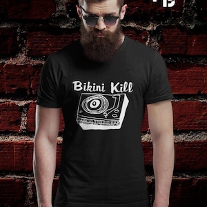 May include: Black t-shirt featuring the words "Bikini Kill" above a white record player graphic. The person wearing the shirt has a beard and sunglasses, set against a red brick wall.