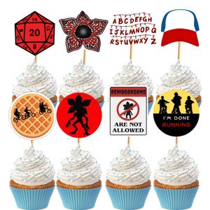 Puede incluir: Cupcakes con adornos temáticos. Los adornos incluyen un dado rojo de 20 caras, una flor Demogorgon, una guirnalda de luces con letras, una gorra de béisbol, un gofre con siluetas y letreros con inscripciones.
