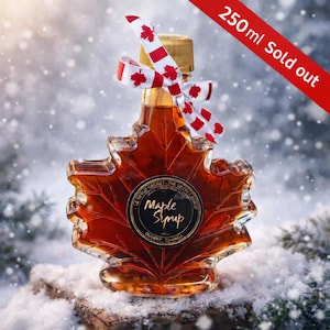 Puede incluir: Una botella de jarabe de arce con forma de hoja de arce, llena de jarabe ámbar. La botella tiene una tapa dorada y está atada con una cinta blanca con diseños de hojas de arce rojas. La etiqueta dice "Maple Syrup, Quebec, Canada". El texto "250ml Sold out" es visible.