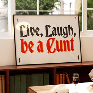 May include: A framed poster with the words "Live, Laugh, be a Cunt" in black and red lettering. The poster is displayed on a wooden shelf with books and a glass of wine. The background includes a window and a view of the outside.
