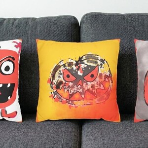 Puede incluir: Tres cojines cuadrados con ilustraciones de calabazas con temática de Halloween. El cojín izquierdo es blanco con una calabaza roja, el central es amarillo anaranjado con una calabaza desgastada y el derecho es gris con una calabaza roja.