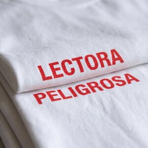 May include: A white t-shirt featuring the red text LECTORA PELIGROSA printed in a bold, sans-serif font. The garment is folded, showing the text clearly across the fabric surface, with the word LECTORA on the top fold and PELIGROSA below.