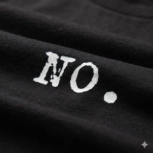 Puede incluir: Primer plano de tela negra con la palabra "NO." impresa en blanco, con letras ligeramente desgastadas. La tela tiene textura y pliegues visibles, creando sombras y profundidad. El punto después de "NO" también es blanco.